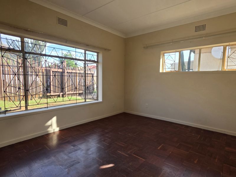 A spacious, empty bedroom featuring original parquet flooring, large windows with security bars, and neutral wall finishes.