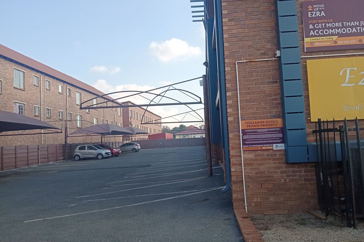 Exterior view of the property featuring a brick facade, paved parking area with shade cloth carports, and perimeter fencing.