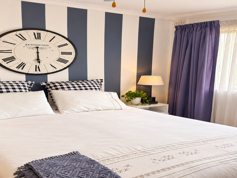 A well-maintained bedroom featuring striped accent wall, large window with layered curtains, and ceiling fan.