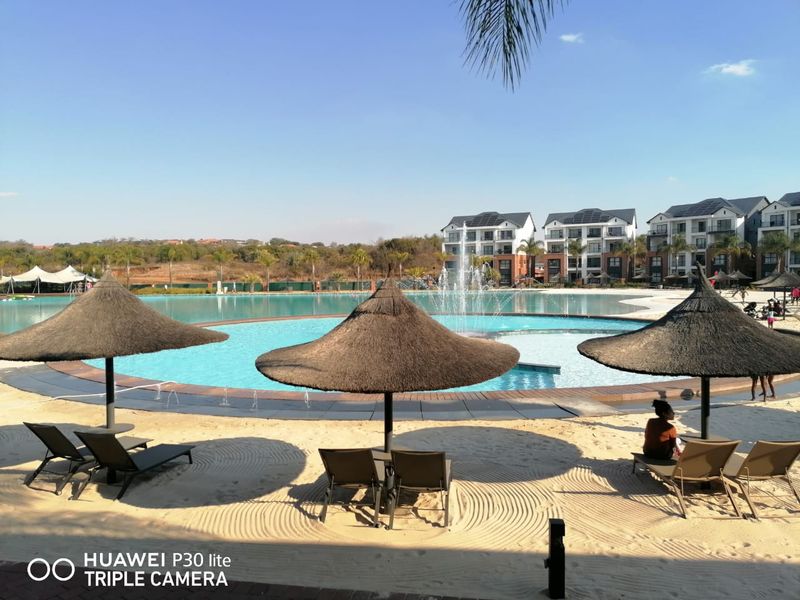A large communal swimming pool area featuring a sandy beach, water fountain, and thatched umbrellas, set within a modern apartment complex.