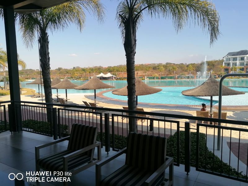A view from a covered patio overlooking a large communal swimming pool with a fountain and landscaped surroundings.