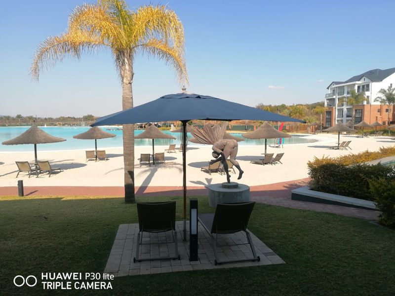 A well-maintained communal pool area featuring a sandy beach-style entry, paved lounging areas, and landscaped gardens within an apartment complex.