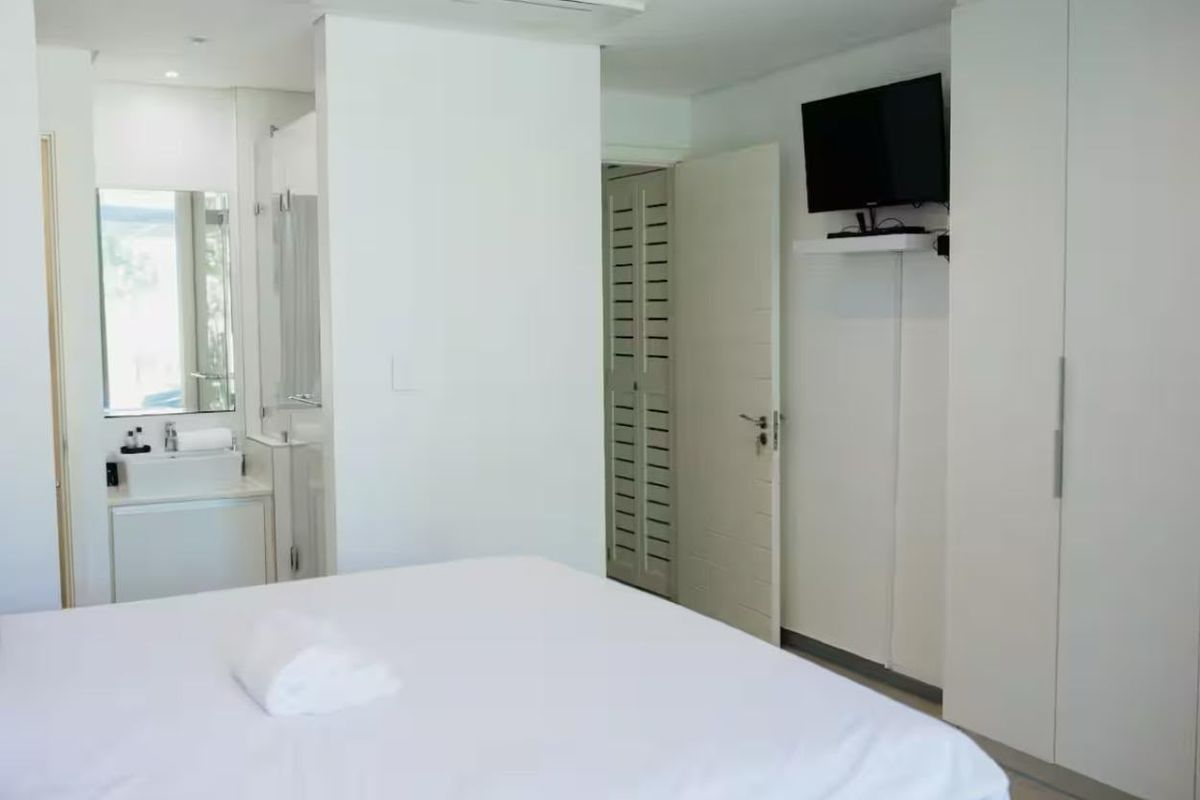 A clean, modern bedroom featuring built-in cupboards and an integrated en-suite bathroom with a vanity basin and shower enclosure.