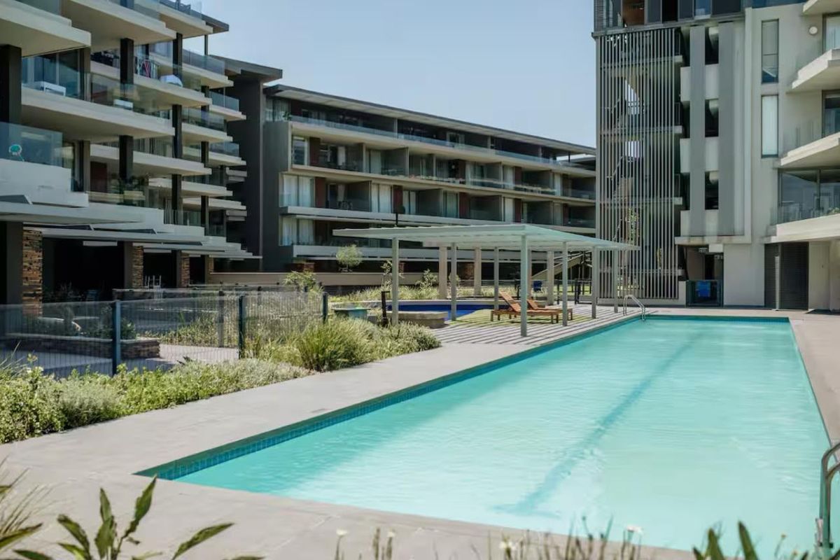 A well-maintained communal swimming pool area featuring a modern pergola, paved surround, and landscaped gardens within a contemporary apartment complex.