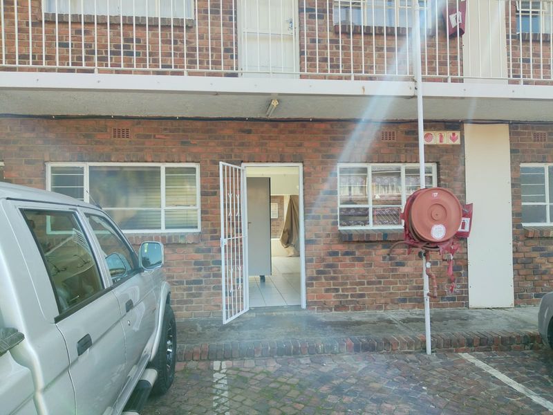Front exterior view of a brick flat featuring a security gate entrance, paved parking area, and building-mounted fire safety equipment.