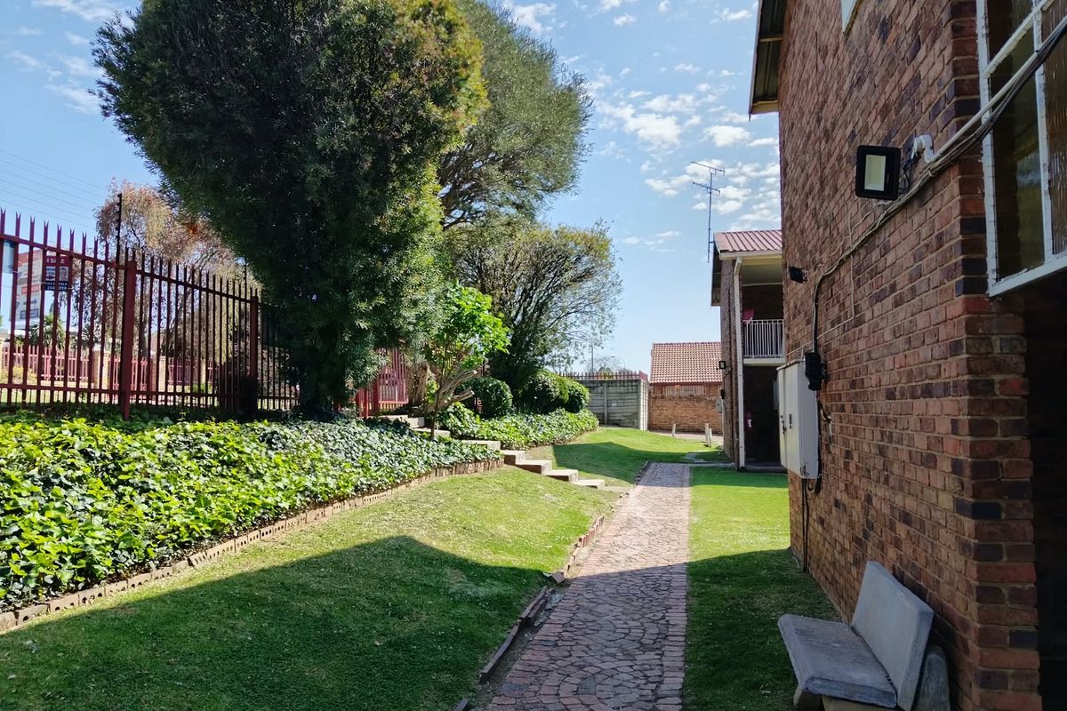 A view of the rear exterior area featuring a brick building facade, a paved walkway, and a well-maintained garden with perimeter fencing.