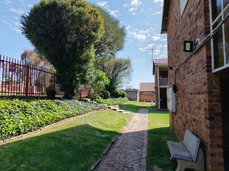 A view of the rear exterior area featuring a brick building facade, a paved walkway, and a well-maintained garden with perimeter fencing.