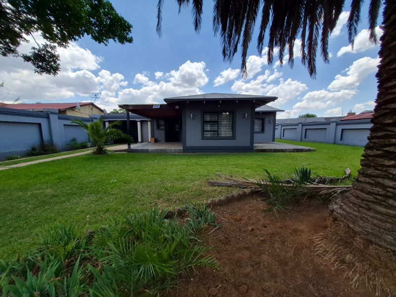 A well-maintained single-story house featuring a modern grey facade, a covered entrance patio, and a secure perimeter wall set within a landscaped garden.