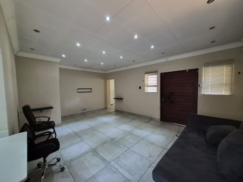 A spacious living area featuring large format floor tiles, recessed downlighting, and a solid timber entrance door with venetian blinds.