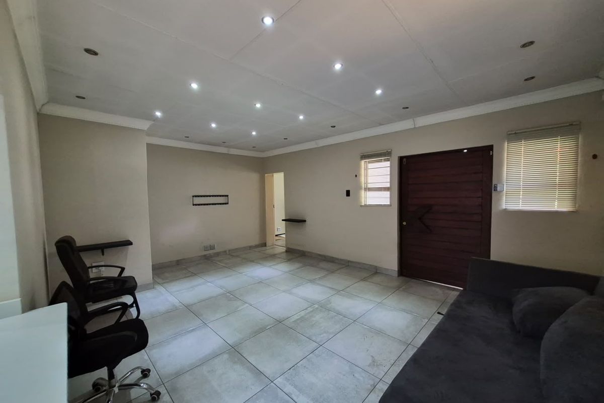 A spacious living area featuring large format floor tiles, recessed downlighting, and a solid timber entrance door with venetian blinds.
