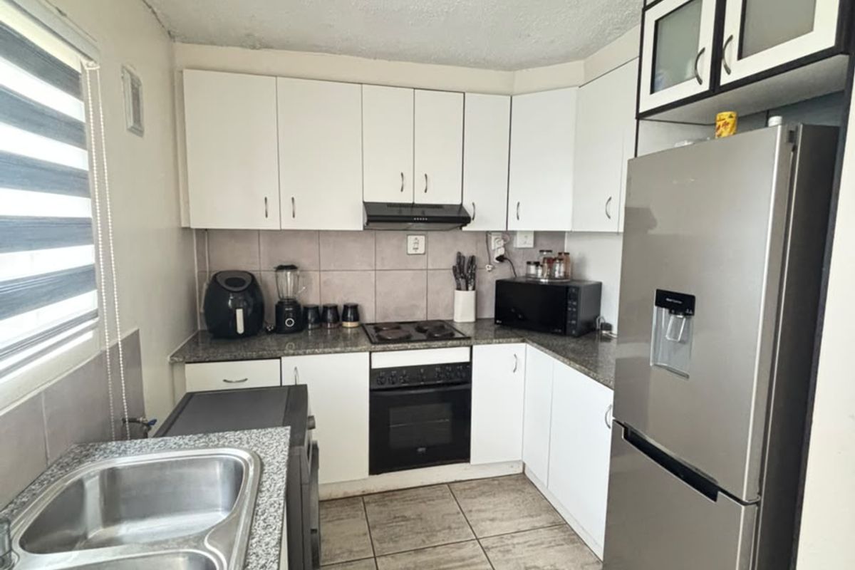 Compact kitchen featuring white cabinetry, granite countertops, built-in oven with hob, and double stainless steel sink, requiring some cosmetic ceiling and flooring repairs.