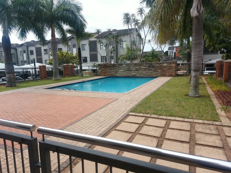 A well-maintained communal swimming pool area featuring a stone feature wall, paved surround, and landscaped garden within an apartment complex.