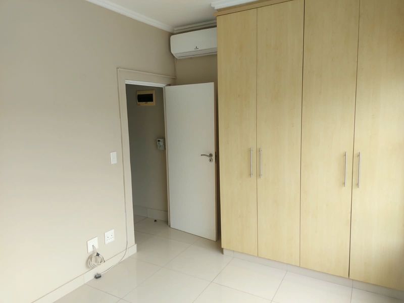 A clean, empty bedroom featuring light-toned built-in cupboards, tiled flooring, and a wall-mounted air conditioning unit.