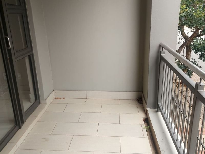 A clean, covered balcony featuring neutral floor tiles and a secure metal balustrade, offering a simple outdoor space accessible via a sliding glass door.