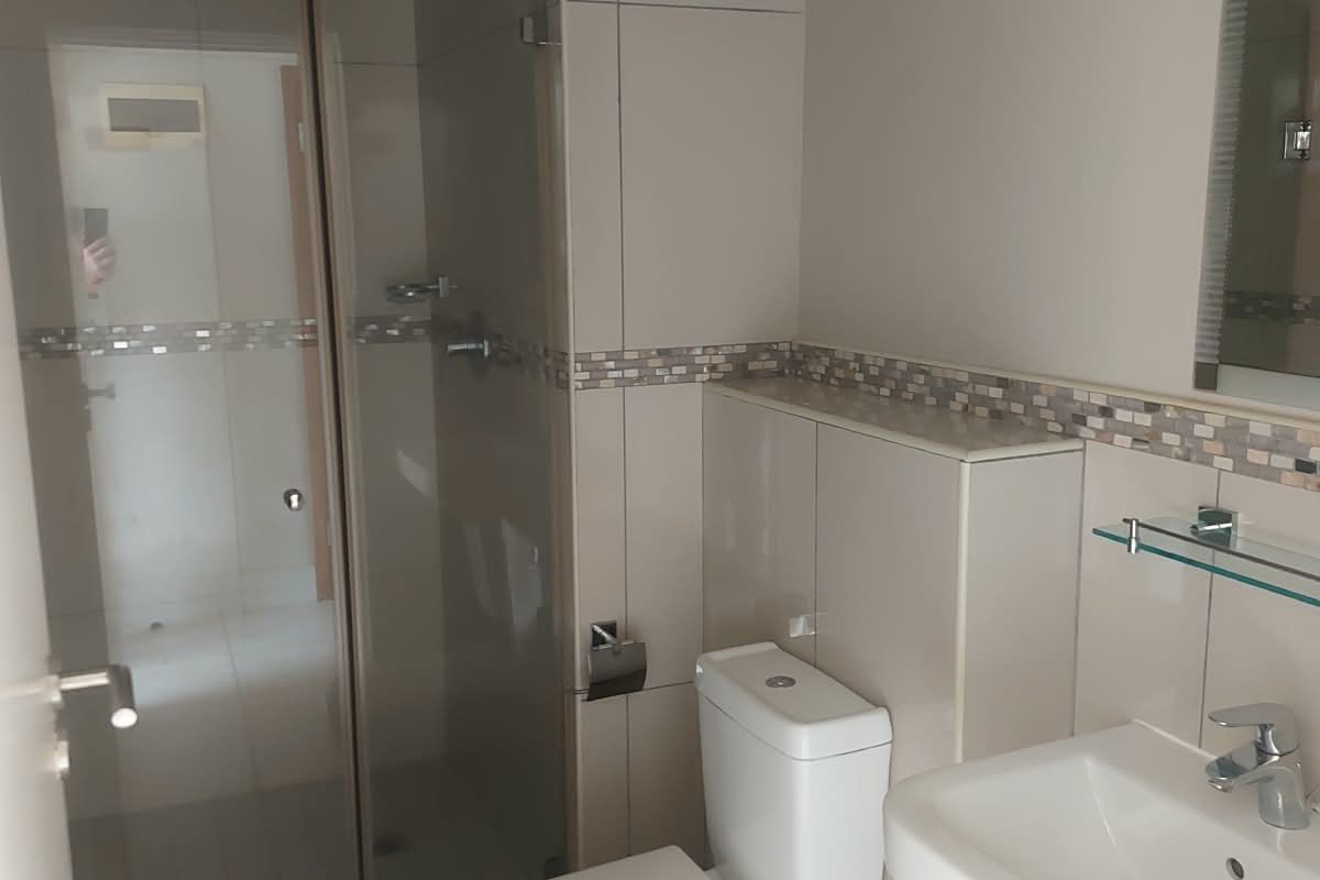 A clean, modern bathroom featuring a glass-enclosed shower, pedestal basin, toilet, and neutral tiled finishes with a decorative mosaic border.
