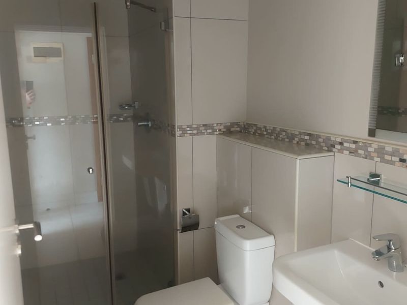 A clean, modern bathroom featuring a glass-enclosed shower, pedestal basin, toilet, and neutral tiled finishes with a decorative mosaic border.