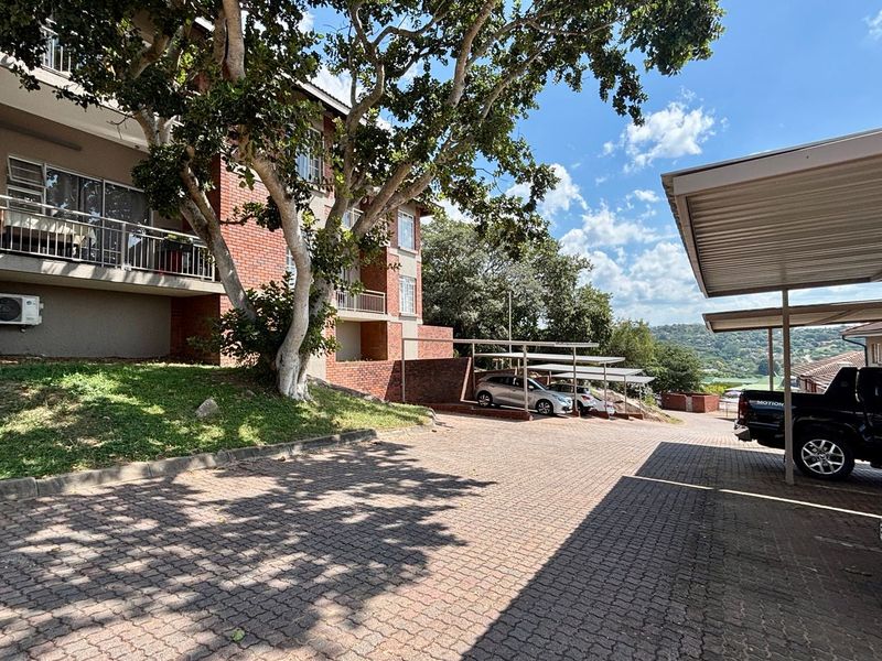 Exterior view of the apartment complex featuring a brick facade, private balconies, paved driveway, and covered carport parking facilities.