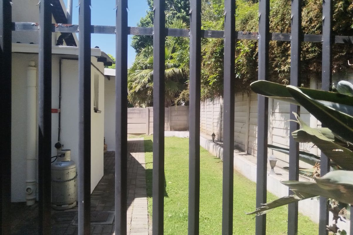 A view of the rear garden area featuring a paved walkway, lawn, and precast concrete boundary wall, viewed through a metal security fence.