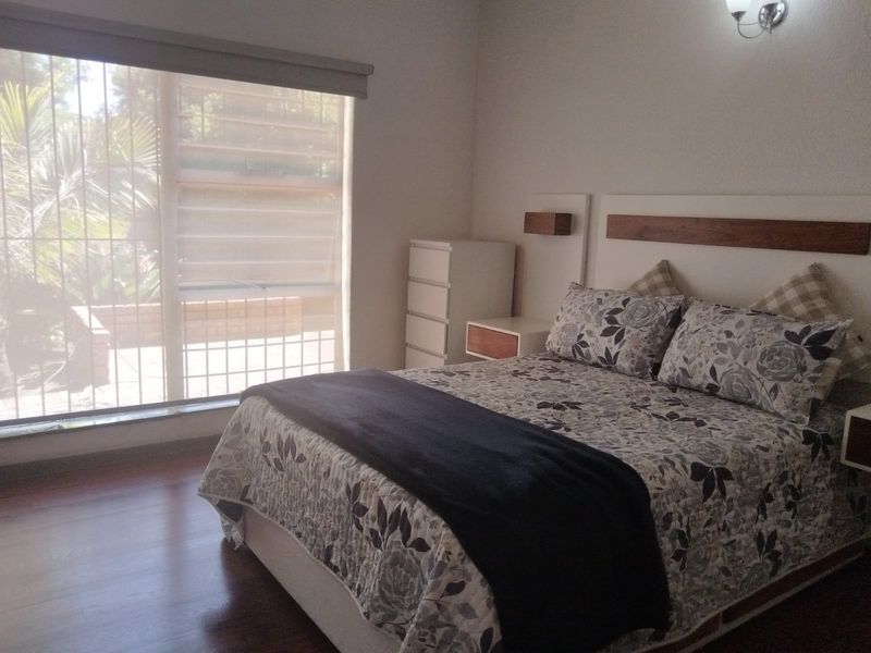 A well-lit bedroom featuring laminate flooring, a custom built-in headboard with integrated bedside tables, and large windows with security bars.
