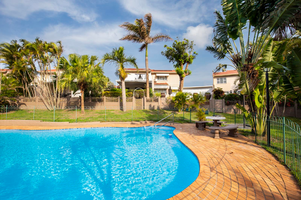 A well-maintained outdoor swimming pool area featuring a paved deck, metal fencing, and established garden landscaping within a townhouse complex.