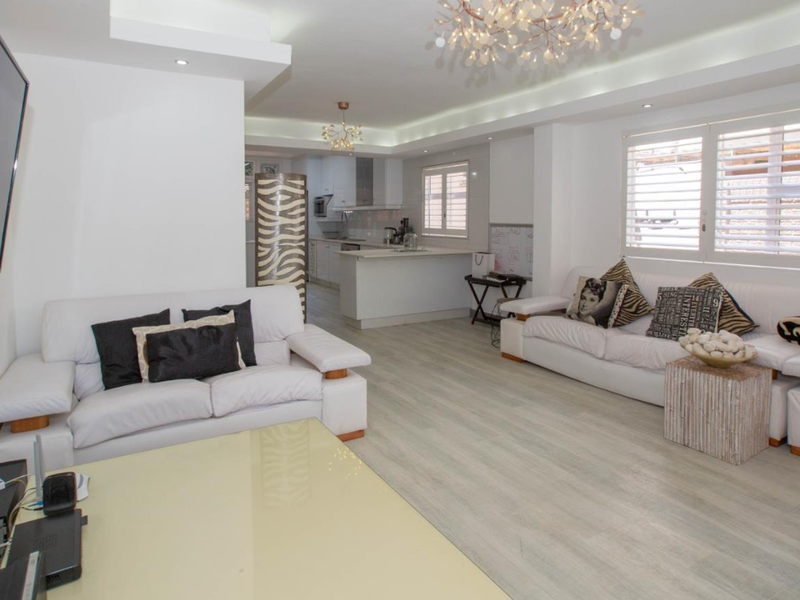 Spacious open-plan living area featuring modern laminate flooring, recessed lighting, and plantation shutters, seamlessly flowing into a well-appointed kitchen with a central island.