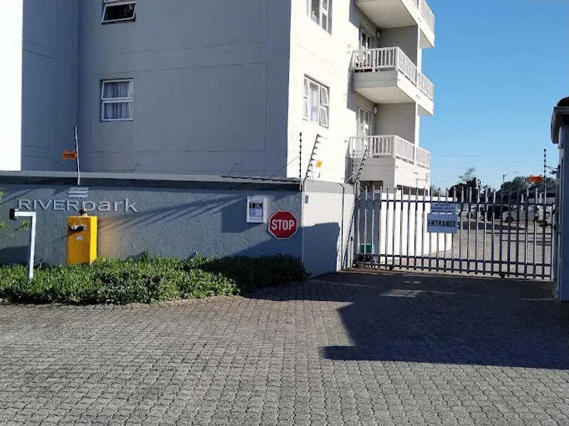 The entrance to Riverpark apartment complex featuring a secure gated access point, paved driveway, and perimeter boundary wall with electric fencing.