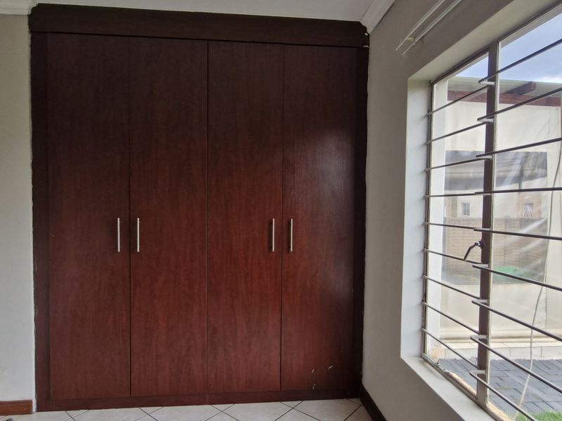 A bedroom featuring built-in wooden cupboards, tiled flooring, and large windows with security bars, offering a functional and neutral space.