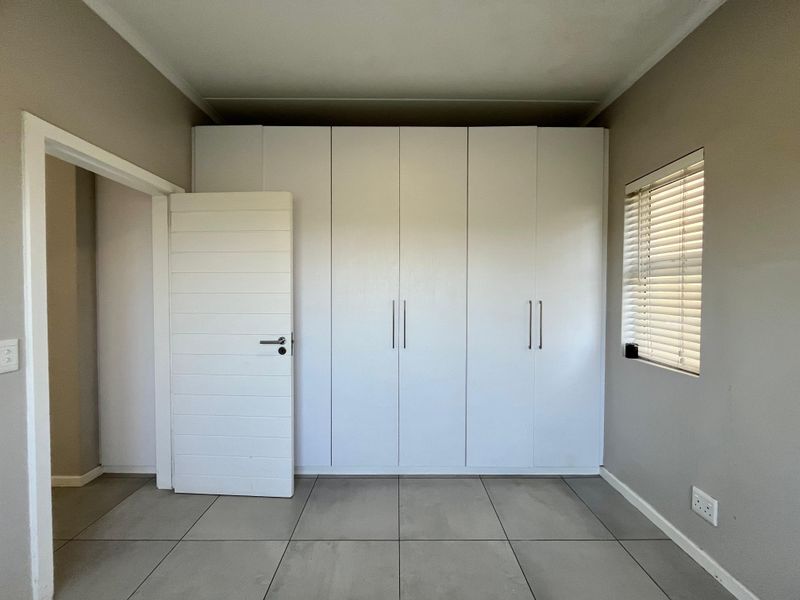 A clean, empty bedroom featuring neutral tiled flooring, built-in white cupboards, and a window fitted with venetian blinds.