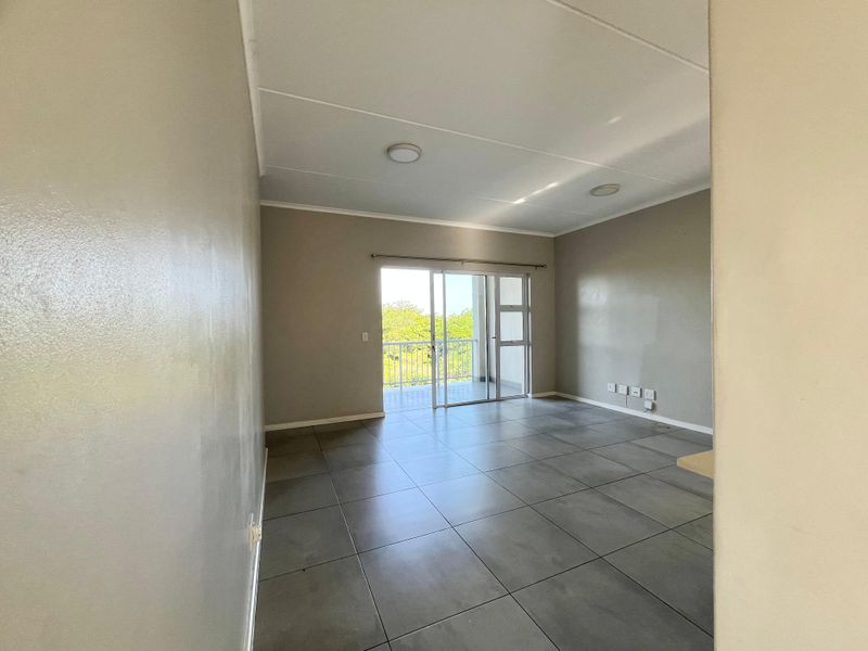 A bright, empty living area featuring large floor tiles, neutral walls, and a sliding glass door providing direct access to an outdoor balcony.
