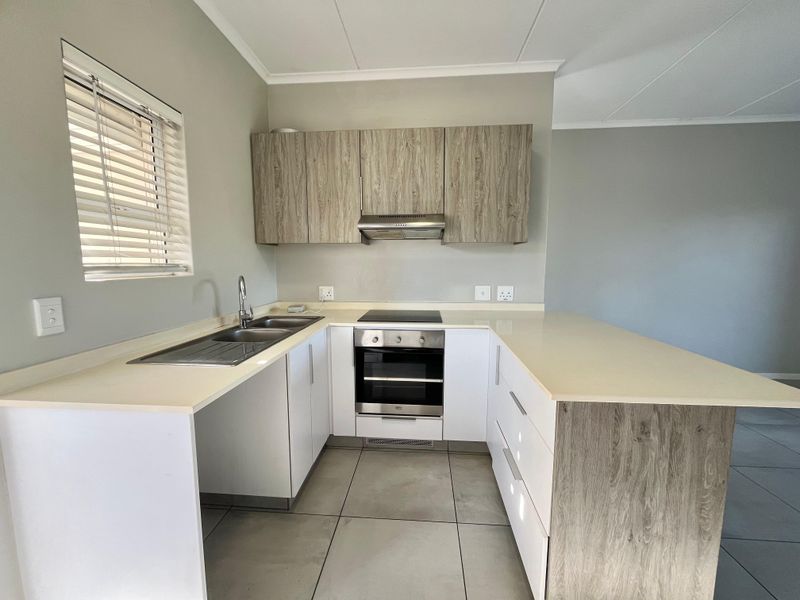 A modern, clean kitchen featuring white and wood-grain cabinetry, stone countertops, integrated oven and hob, and tiled flooring in an open-plan layout.
