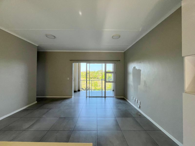 An empty, open-plan living area featuring large grey floor tiles, neutral walls, and a sliding glass door leading to a balcony with outdoor views.