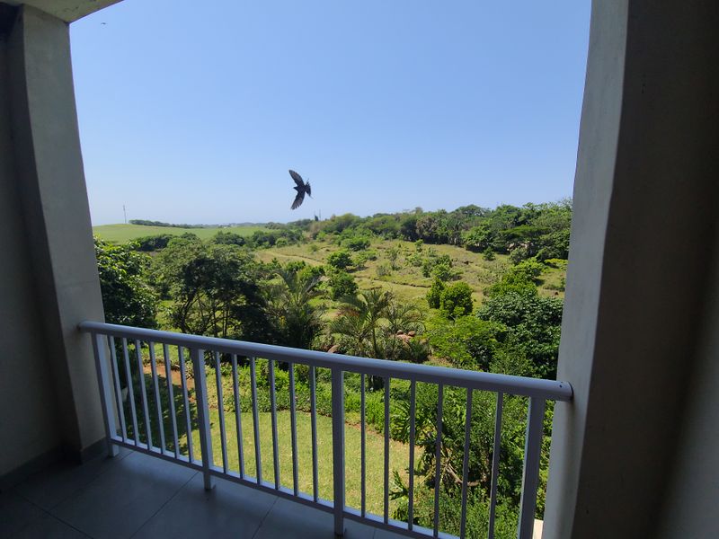 A covered balcony featuring a white metal balustrade and tiled flooring, offering an expansive view of the surrounding green landscape.