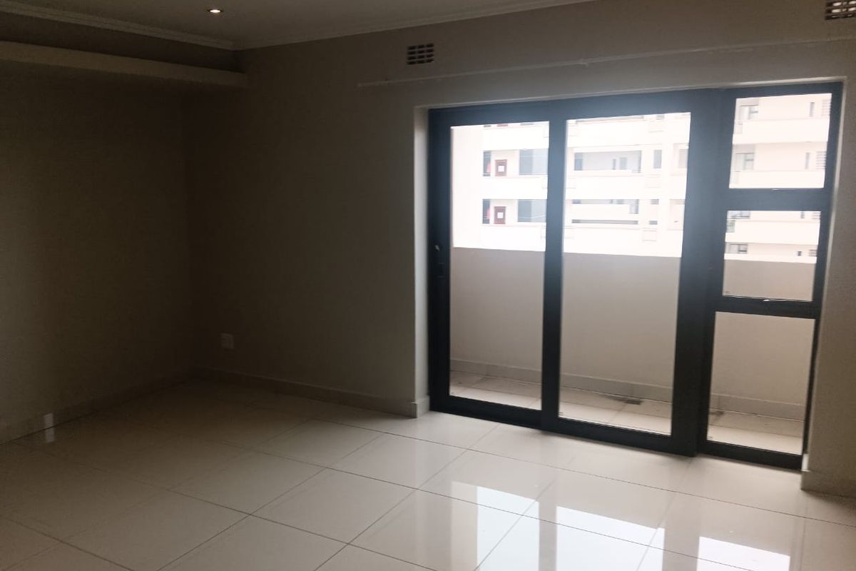 A bright, empty living area featuring neutral tiled flooring and a large sliding glass door providing access to an outdoor balcony.