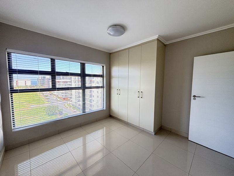 A bright, empty bedroom featuring neutral tiled flooring, built-in cupboards, and large windows with venetian blinds offering an exterior view.