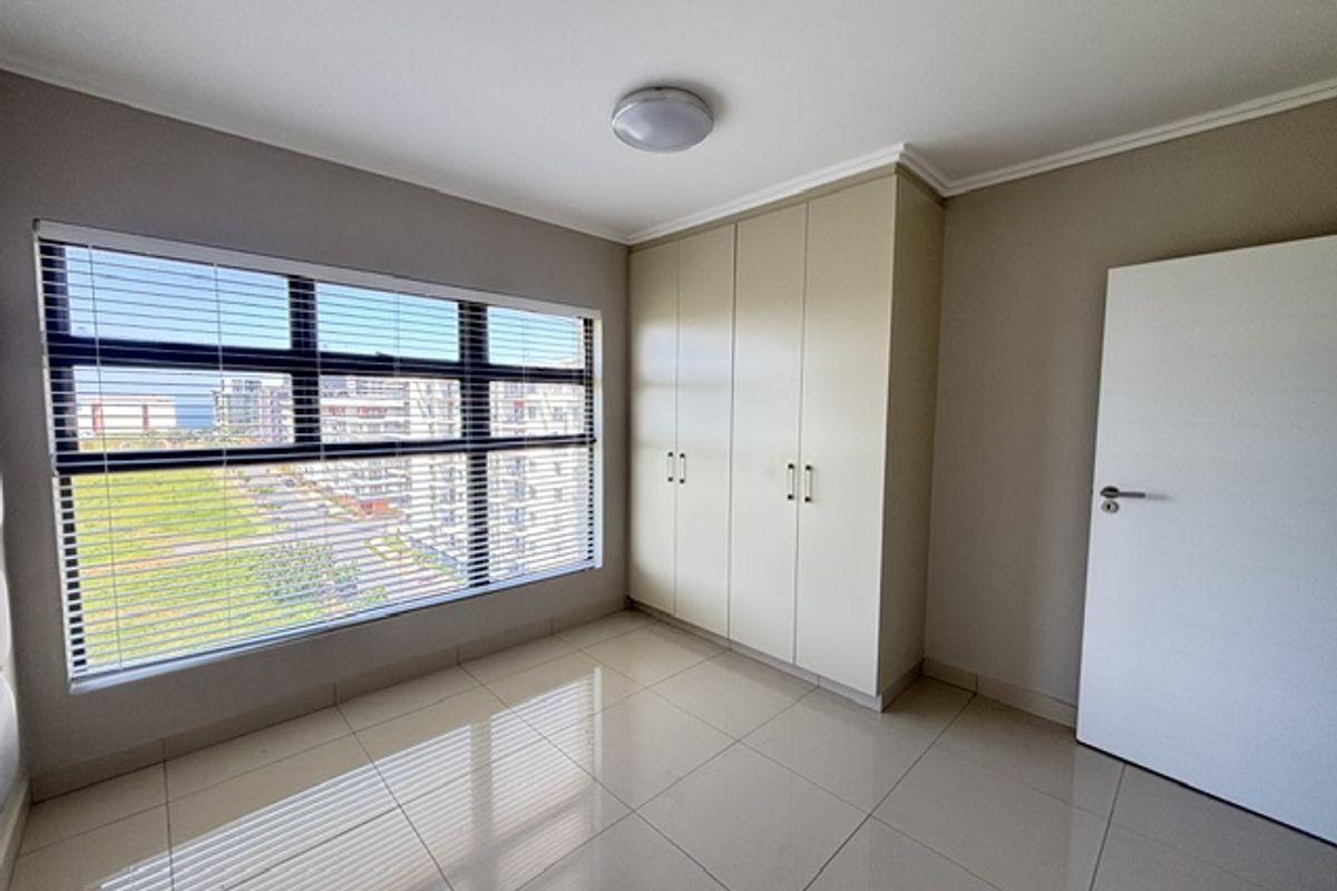 A bright, empty bedroom featuring neutral tiled flooring, built-in cupboards, and large windows with venetian blinds offering an exterior view.