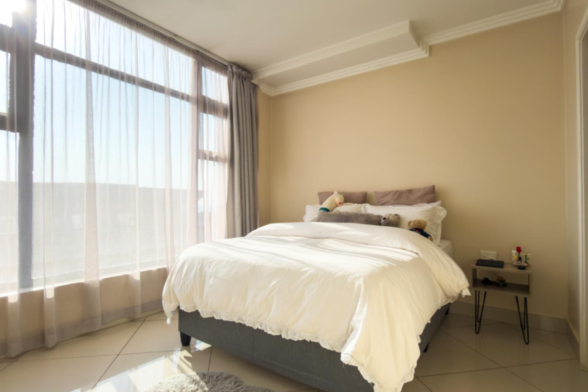 A bright and airy bedroom featuring large floor-to-ceiling windows that allow for ample natural light, complemented by neutral tiled flooring and coved ceiling details.