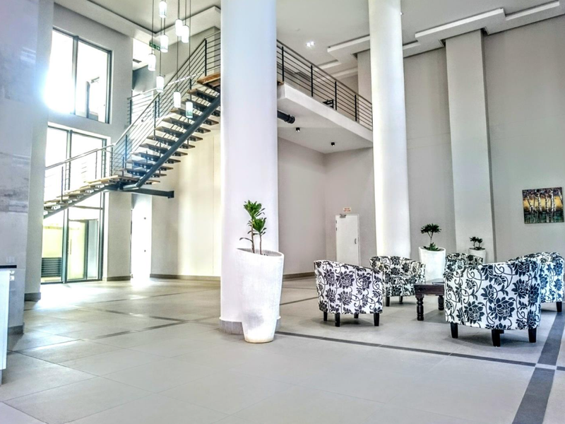 Spacious modern apartment building lobby featuring high ceilings, large structural columns, tiled flooring, and an open-plan staircase with metal railings.