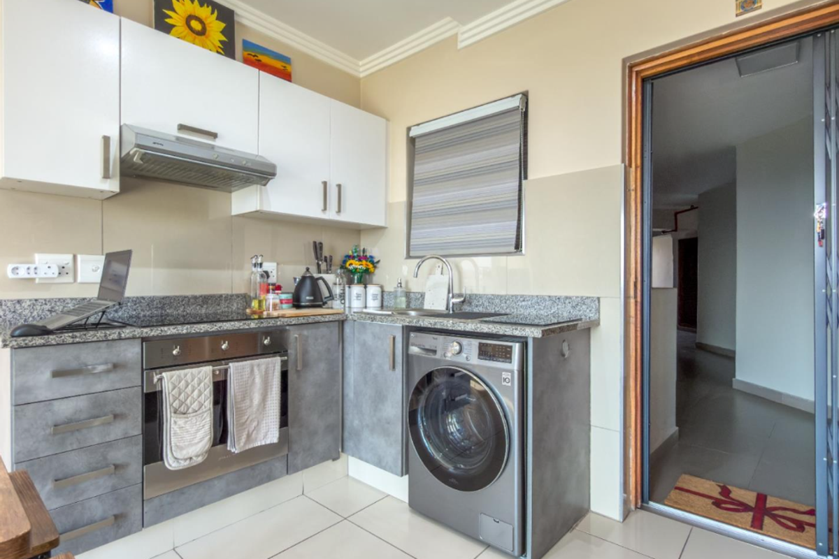 A modern, compact kitchen featuring granite countertops, built-in cabinetry, a stainless steel oven, and an integrated washing machine, finished with neutral tiled flooring.