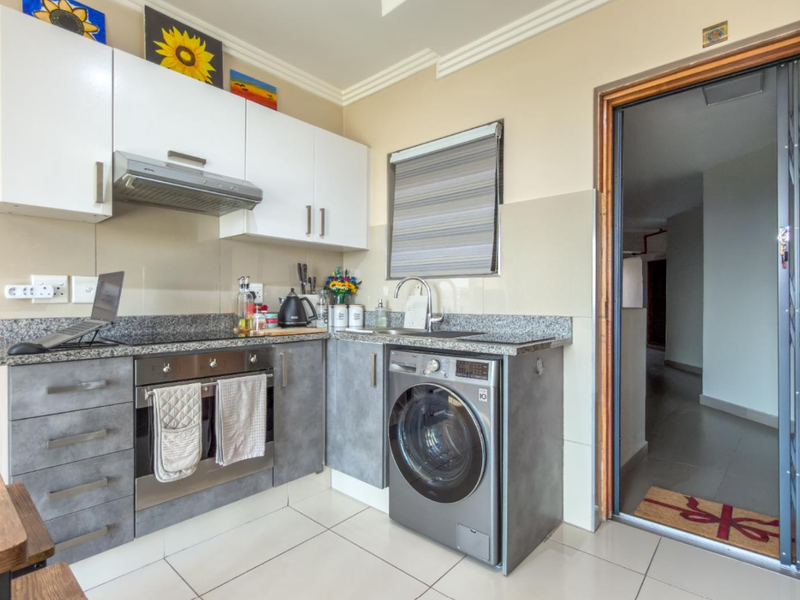 A modern, compact kitchen featuring granite countertops, built-in cabinetry, a stainless steel oven, and an integrated washing machine, finished with neutral tiled flooring.