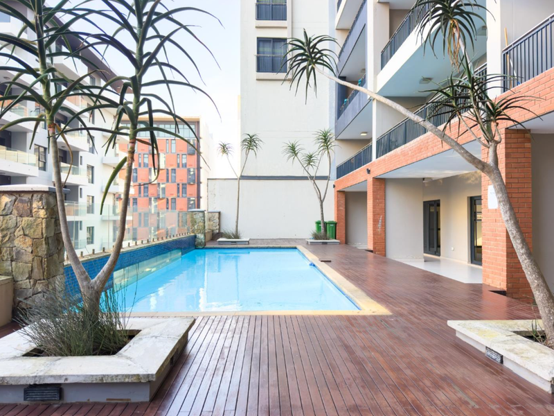 Communal swimming pool area featuring timber decking, brick structural pillars, and glass balustrades within an apartment complex setting.