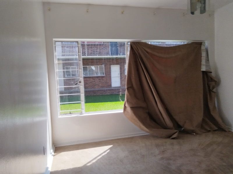 A vacant bedroom featuring carpeted flooring and large windows fitted with security bars, offering a view of the exterior complex grounds.