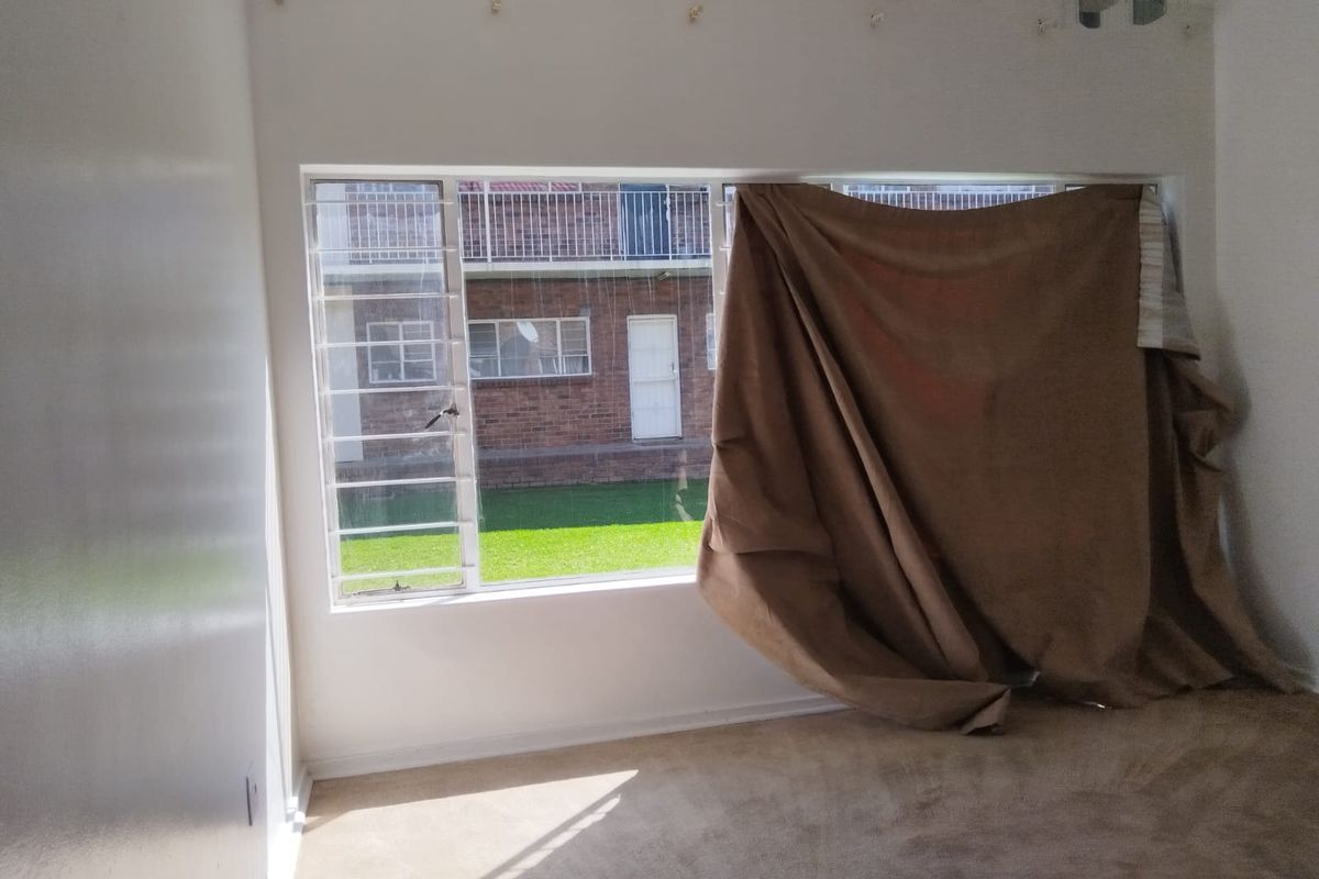 A vacant bedroom featuring carpeted flooring and large windows fitted with security bars, offering a view of the exterior complex grounds.