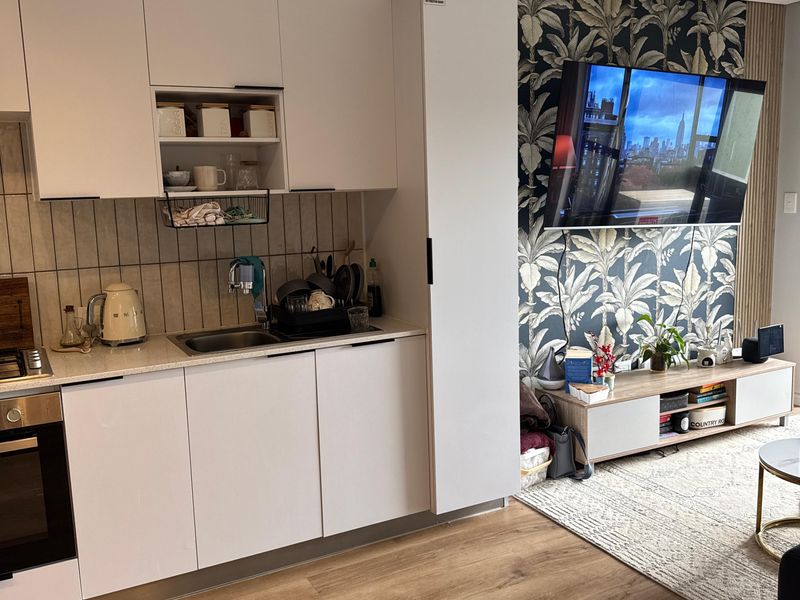 Modern open-plan apartment featuring a compact kitchen with white cabinetry and integrated appliances, seamlessly flowing into a living area with feature wallpaper.