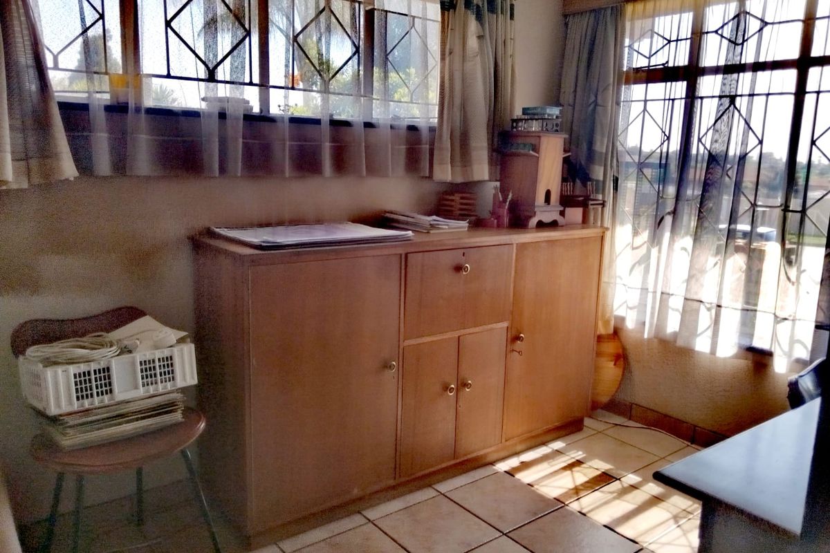 A living area featuring tiled flooring, built-in wooden cabinetry, and large windows with security bars, offering a dated but functional space.