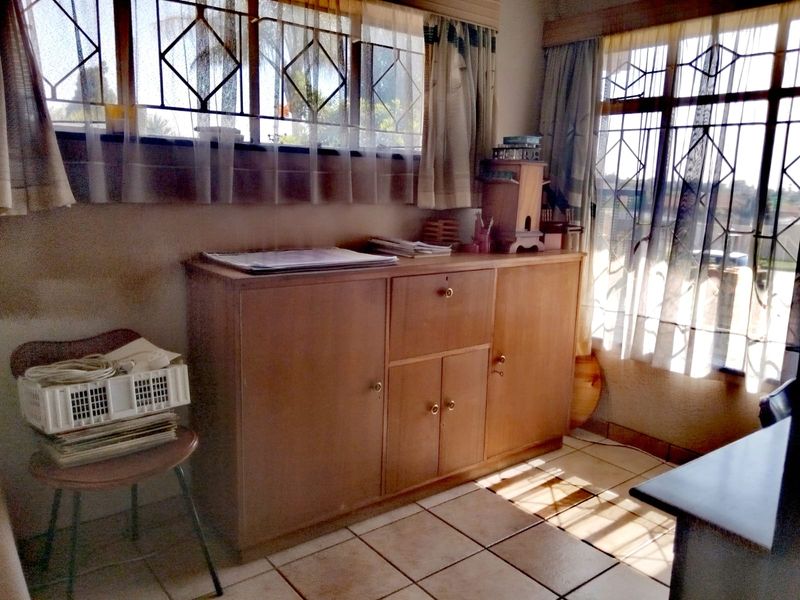 A living area featuring tiled flooring, built-in wooden cabinetry, and large windows with security bars, offering a dated but functional space.