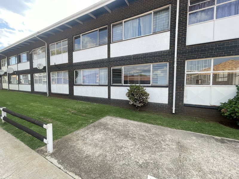 Rear view of a multi-story apartment building featuring a brick facade, aluminium windows with security bars, and a shared lawn area with concrete paving.