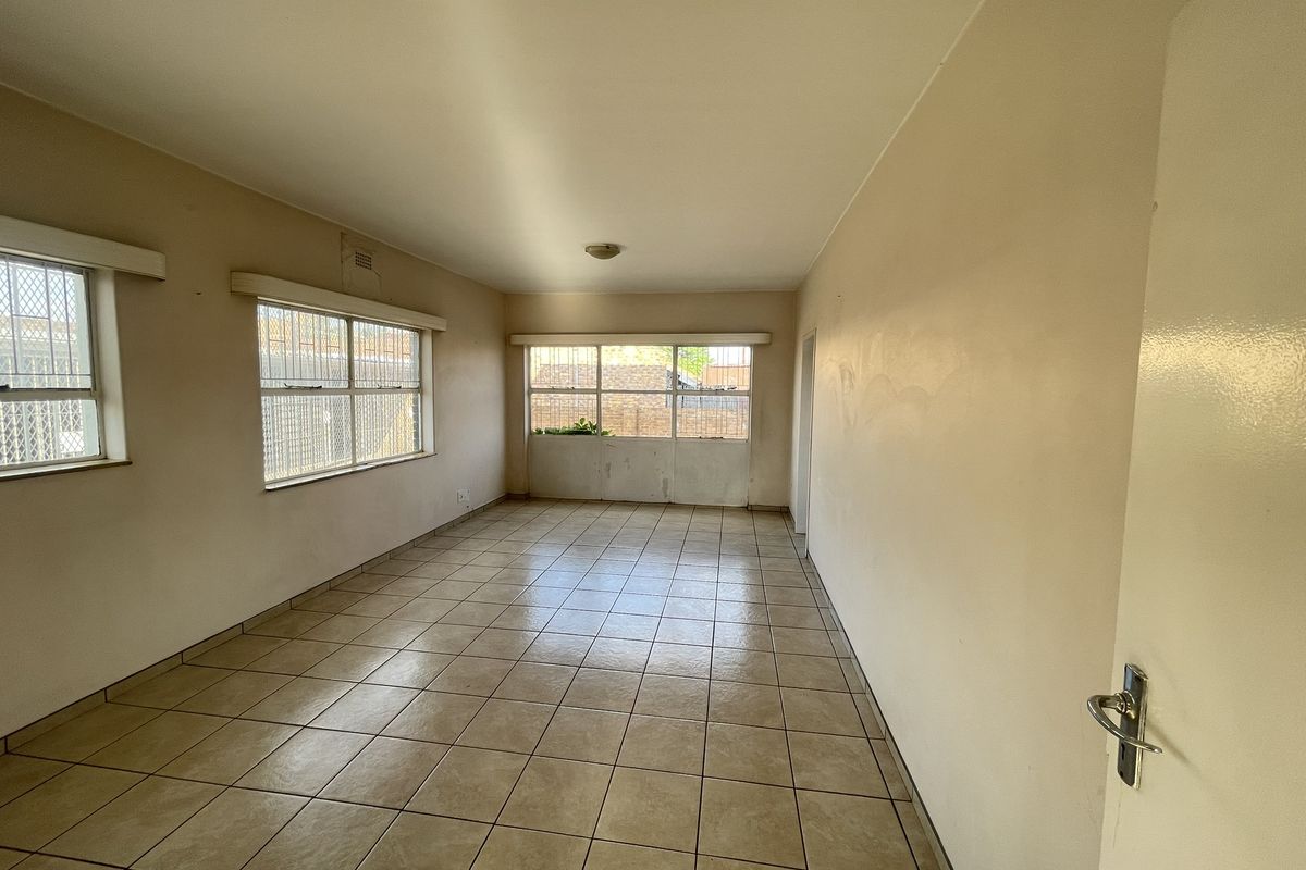 An empty, neutrally painted living area featuring tiled flooring and large windows with security bars, offering a blank canvas for potential renovation.