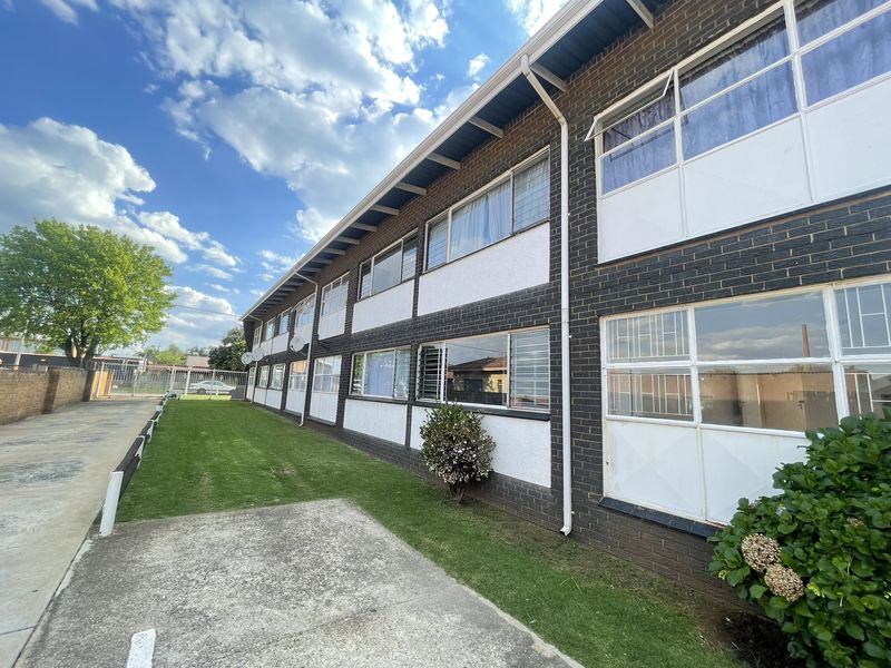 Exterior view of a multi-story apartment building featuring brick and plaster facade, aluminium windows with security bars, and adjacent paved parking area.