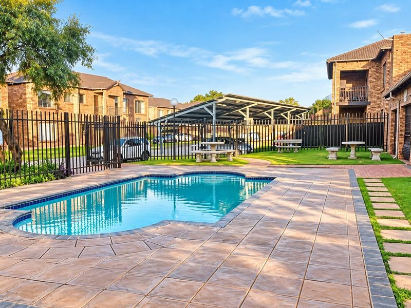 A well-maintained communal pool area featuring a paved deck, metal fencing, and adjacent carport parking within a residential apartment complex.