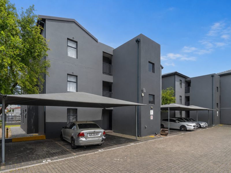 Modern multi-story apartment complex featuring a grey facade, paved parking area, and dedicated shade cloth carports for residents.